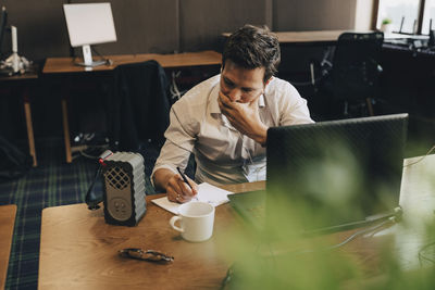 Businessman covering mouth while writing in paper at office desk