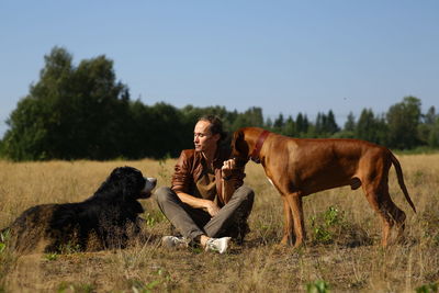 Man with dogs while sitting on grassy land against sky