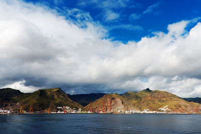 Cranes at harbor in sea by mountains against cloudy sky at canary islands