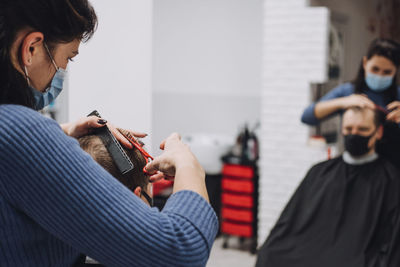 Close-up of female barber in mask cutting hair of man in salon