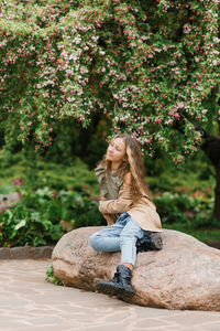 Cute teenage girl with long brown hair in a trench coat is sitting on a rock in the park