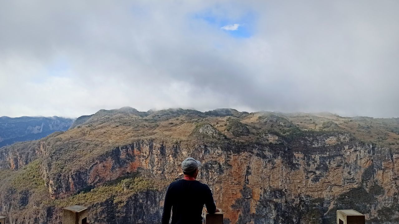 REAR VIEW OF PERSON STANDING ON MOUNTAIN AGAINST SKY