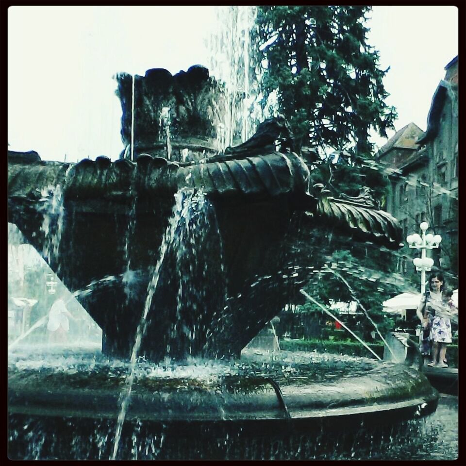 water, transfer print, fountain, splashing, motion, auto post production filter, clear sky, spraying, waterfront, built structure, tree, architecture, building exterior, waterfall, day, flowing water, sunlight, outdoors, long exposure, sky