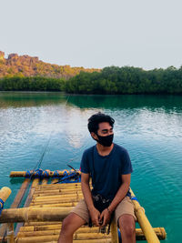 Young man wearing mask sitting on bamboo raft in lake