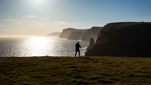 Silhouette man standing on land by sea against sky during sunset