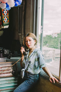 Portrait of young woman talking over telephone while standing by window