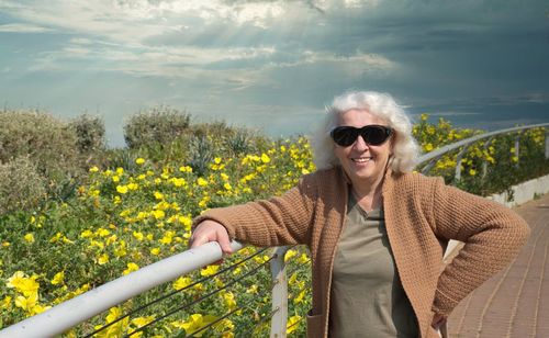 Mature gray-haired happy woman over 60, wearing sunglasses, against the background of yellow flower