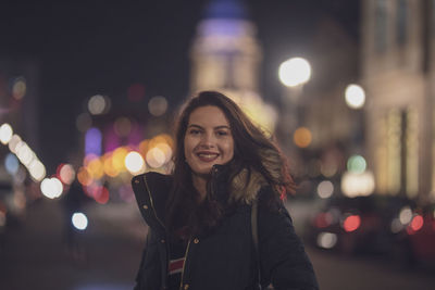 Portrait of young woman smiling while standing in illuminated city at night