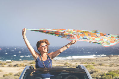 Woman holding scarf while standing in car against sky