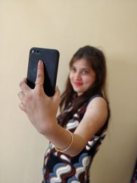 Young woman taking selfie through mobile phone by wall at home