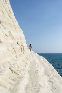 Shirtless man walking on rock by sea against clear blue sky