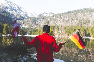 Rear view of mature man holding national flags while standing by lake against mountain