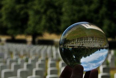 Close-up of hand holding crystal ball with reflection of trees and world war 2 graveyard