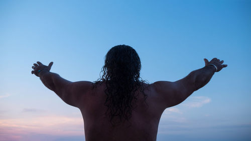 Rear view of shirtless man with arms raised standing against sky during sunset