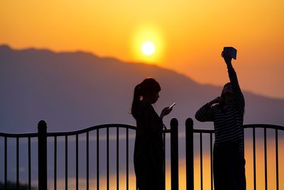 Silhouette people photographing railing against sky during sunset