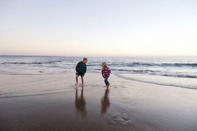 Playful siblings running on shore at beach against sky during sunset