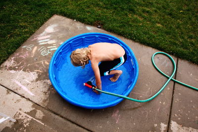 High angle view of boy holding garden hose in wading pool at backyard