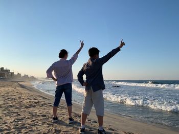 Siblings with hand raised standing at beach against clear sky