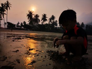 Boy playing with sand against palm trees during sunset