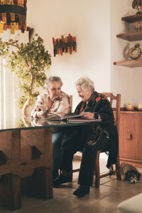 Elderly sisters inspecting pictures in photo album and discussing memories while sitting at table at home together