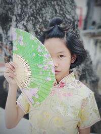 Portrait of cute girl holding folding fan standing against tree trunk