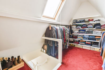 Collection of various elegant suits and outerwear hanging on racks near window in light attic with wooden drawer in apartment