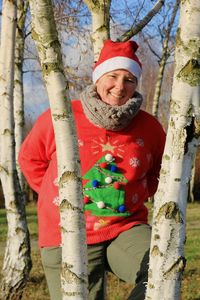 Portrait of smiling woman standing by tree trunk with christmas sweater