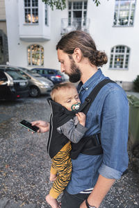 Mid adult man using mobile phone while carrying baby in carrier on sidewalk