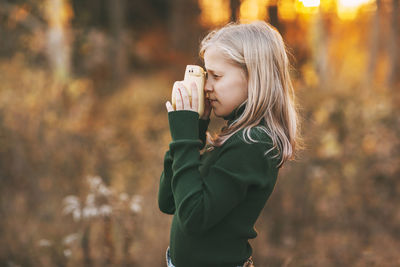 Teenage girl with blond hair takes pictures of nature with a polaroid camera in an autumn park