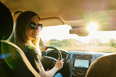 Woman wearing sunglasses is driving car with sunlight through windshield