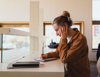 Side view of young woman in casual clothes with hair bun touching head and writing in planner while sitting at table and studying in university library