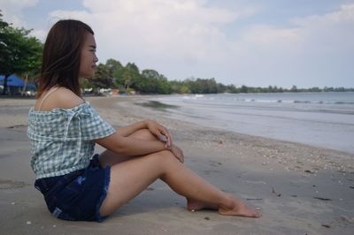 Side view of young woman sitting on beach against sky