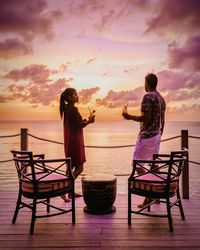 Couple enjoying drinks at pier by sea against sky during sunset
