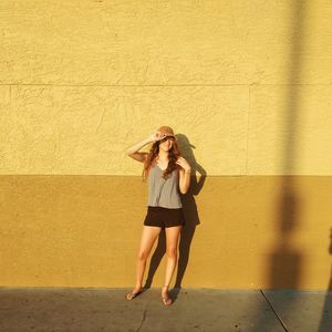 Young woman wearing cap while standing on footpath against yellow wall during sunny day