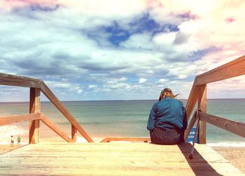 Rear view of woman sitting on steps by sea against cloudy sky