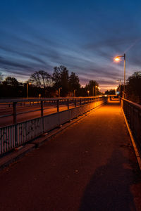 Road by illuminated street against sky at sunset