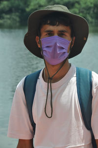 A young male tourist wearing face mask with looking at camera while standing beside of lake