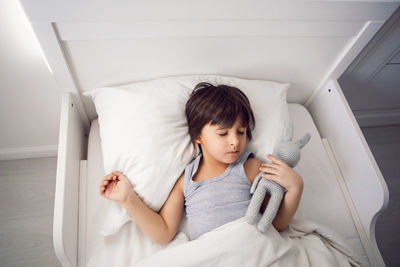 Child boy lies in a white children's bed with a blanket in a room with toys rabbits