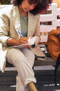 Crop thoughtful african american female entrepreneur in suit sitting on bench and writing in planner while working remotely on sunny day in street restaurant