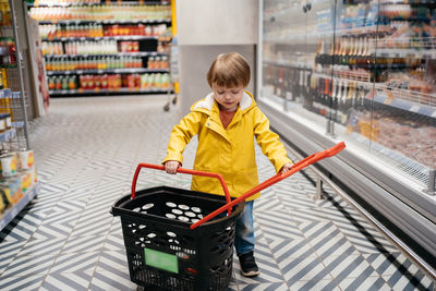 Child in the market with a grocery cart, wearing a yellow jacket and jeans