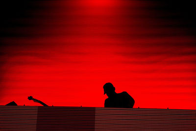 Silhouette man sitting against red wall against sky during sunset