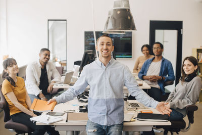 Portrait of smiling businessman gesturing while standing with colleagues sitting at desk in creative office