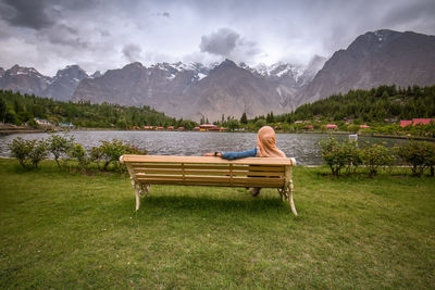 Woman sitting on bench by lake against mountains