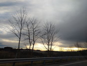 Silhouette bare trees by road against sky during sunset