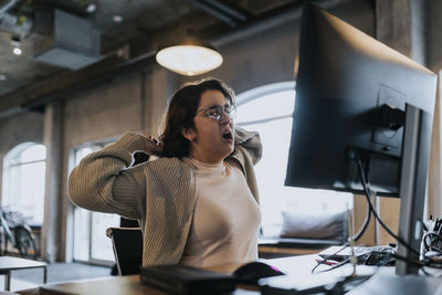 Tired female hacker yawning and stretching hands at desk in creative office