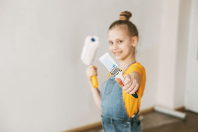 A girl in a denim overalls and a yellow t-shirt helps to paint the walls in an apartment white.