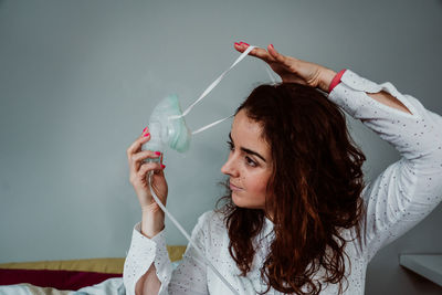 Woman wearing oxygen mask while sitting on bed against wall in hospital