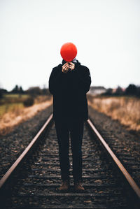 Man covering face with red balloon while standing on railroad track against sky