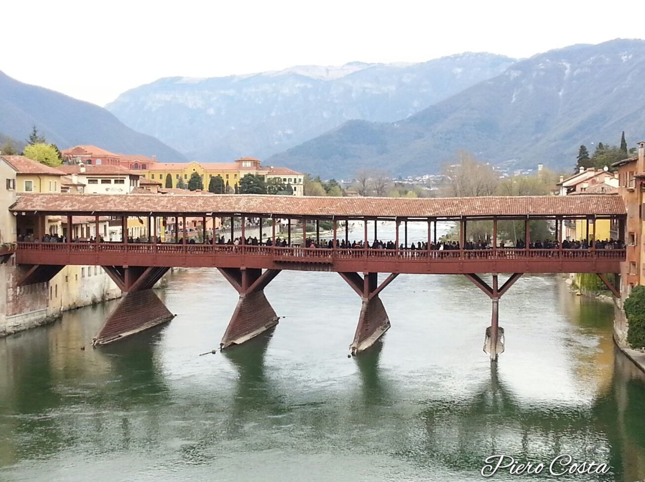 mountain, mountain range, water, built structure, lake, waterfront, architecture, river, tranquility, nature, scenics, tranquil scene, reflection, beauty in nature, day, building exterior, outdoors, clear sky, bridge - man made structure, sky