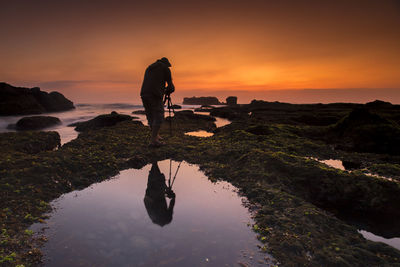 Silhouette man photographing while standing on rock by sea against sky during sunset